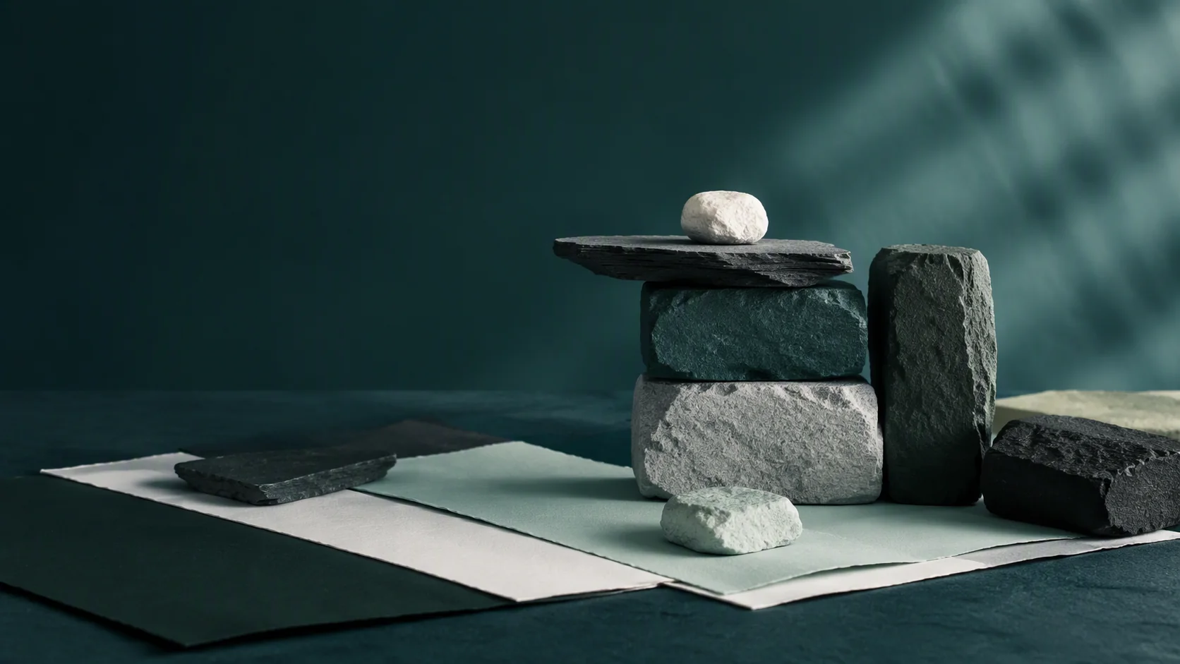 An abstract arrangement of balanced stone blocks and layered teal paper-like planes in a quiet landscape.